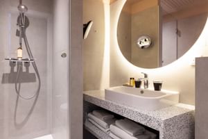 Zeitgenössisches Badezimmer mit Glasdusche, weißem Waschbecken auf gesprenkeltem Steinwaschtisch, hinterleuchteter runder Spiegel.