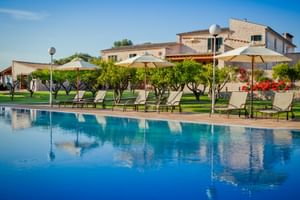 Großer blauer Pool mit Sonnenliegen und Sonnenschirmen in der Finca Son Roig. Steingebäude und gepflegte Gärten mit Bäumen im Hintergrund.