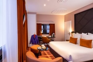 Elegantes Hotelzimmer mit großem Bett, dunklem gestepptem Kopfteil, orangefarbenen Kissen und Sitzbereich mit orangefarbenen Stühlen.