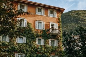 Orange four-story building with white shutters and ivy-covered walls, balcony on second floor, surrounded by trees with mountains in background.