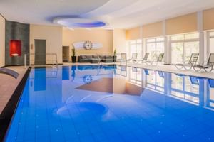 Hallenbad im Gartenhotel Heusser Bad Dürkheim mit blauem Wasser, Liegestühlen, großen Fenstern und dekorativer Decke mit blauer Beleuchtung.