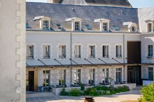 White stone building of Hotel Fleur de Loire in Blois with gray slate roof, dormer windows, and terrace with awnings facing a sunny courtyard.