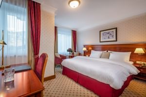 Elegantes Hotelzimmer mit Doppelbett, roten Polstern, gemustertem Teppich, Holzmöbeln, zwei Fenstern mit roten Vorhängen und klassischem Dekor.