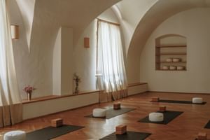 Vaulted yoga studio with yoga mats, cushions, and wooden blocks on parquet floor. Arched ceiling, white curtains, and wall niches create a calm space.