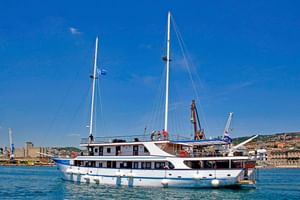 White motor yacht MS Aria with two masts and blue trim moored in harbor with white fenders. Coastal town visible in background under clear blue sky.