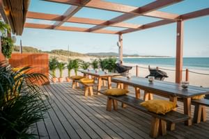 Wooden deck terrace with benches and tables under pergola overlooking Cabedelo Beach. Palm plants frame the view of sandy beach and ocean.