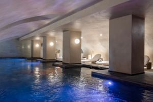 Modern indoor spa pool with blue lighting at Hotel Fleur de Loire Blois, featuring white columns, lounge chairs, and ambient ceiling lighting.
