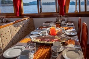 Breakfast table on MS Macek with plates, cups, juice, and cold cuts. Large windows with red curtains show lake view with forested shores.