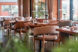 Modern restaurant interior with wooden tables, beige upholstered chairs, and large windows. Tables are set with glasses and candles.