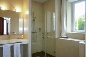 Helles Bad mit beigen Fliesen, Glasdusche, weißem Waschbecken, Spiegel mit Wandleuchten, Badewanne am Fenster im Le Grand Monarque.