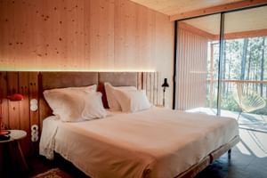 Double room with white bedding and beige headboard in FeelViana Sport Hotel. Wood-paneled walls and glass doors open to a balcony with forest views.