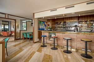 Modern hotel bar with marble counter, colorful bar stools, and espresso machine. Striped marble floor leads to lounge area with turquoise chairs.