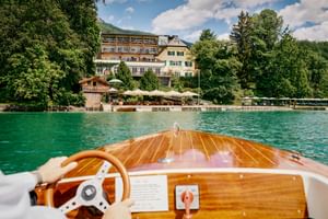 View from wooden boat approaching Landhaus zu Appesbach hotel on turquoise lake. Hotel buildings with terrace and umbrellas visible on shore.