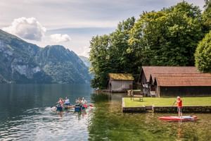 Menschen beim Kajakfahren und Stand-up-Paddeln am Traunsee mit traditionellen Holzbootshäusern und Bergen im Hintergrund bei Traunkirchen.