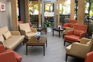 Modern hotel lounge with beige and coral armchairs around dark wood tables. Natural wood pillars and glass doors open to garden terrace.