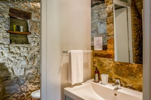 Modern bathroom at Le Valli Agriturismo with white sink, mirror, and exposed stone walls. Wooden beam visible above sink with towel rail.