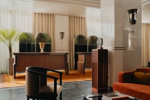 Art-Déco-Empfangsbereich im The Dean München mit dunklen Holzmöbeln, orangefarbenem Samtsofa, gewölbten Spiegeln und beigen Vorhängen.