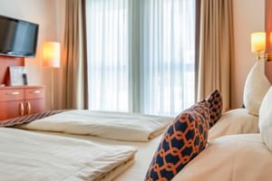 Helles Doppelzimmer mit zwei Betten, Zierkissen, TV, Stehlampe, Wandlampe und großen Fenstern mit transparenten und beigen Vorhängen.