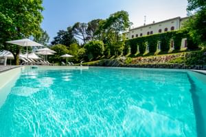 Türkisfarbener Pool der Villa Soligo in Farra di Soligo mit weißen Sonnenliegen, umgeben von grünen Bäumen und efeubedecktem Hang.