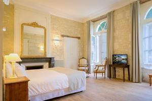 Elegantes Doppelzimmer mit weißer Bettwäsche, Steinwänden, goldenem Zierspiegel über Kamin, hohen Bogenfenstern mit beigen Vorhängen und klassischen Möbeln.