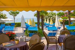 Terrace with dining tables under yellow awning at Du Lac et Bellevue hotel in Bardolino, overlooking Lake Garda with blue loungers and parasols.