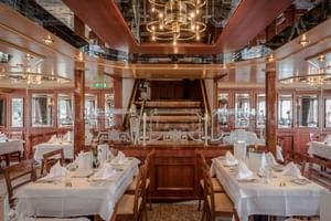 Elegant dining room on MS Switzerland with white-clothed tables, wooden chairs, and a central wooden staircase under an ornate chandelier.