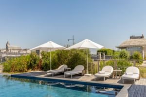Rooftop pool with wooden deck, white sun loungers, and umbrellas overlooking Blois cityscape under clear blue sky.