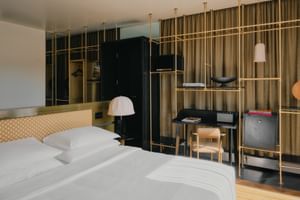 Zeitgenössisches Hotelzimmer im The Dean München mit weißem Bett, goldenem Regalsystem, Schreibtisch und beigen Vorhängen.
