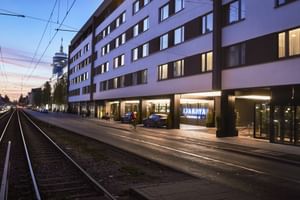 Moderne weiße Fassade des The Dean Munich Hotels bei Dämmerung mit beleuchteten Eingang und Fenstern. Straßenbahngleise verlaufen entlang der Straße.