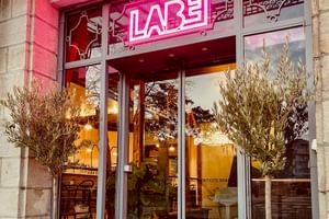 Modern glass entrance of Hotel Labé with pink neon signage above the door. Two olive trees in white pots flank the entrance.