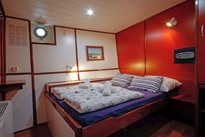 Double cabin on MS Macek with wooden bed, striped pillows, porthole window, and warm wood paneling. A safe is mounted on the red wall.