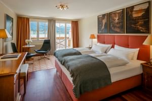 Spacious hotel room with two beds, coral-colored headboards, dark wood floors, mountain view through balcony doors, and orange curtains.
