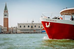 Rot-weißes Schiff MS Ave Maria in der Lagune von Venedig mit Campanile und Dogenpalast im Hintergrund unter klarem Himmel.