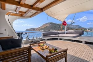 Oberdeck der Yacht Boreas mit grauer Sitzecke, Holzcouchtisch mit Speisen, Teakdeck und Küstenbergblick im Hintergrund.
