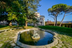 Runder Steinbrunnen mit Skulptur im Garten der Villa Soligo, Farra di Soligo. Weiße Villa und hohe Pinien unter blauem Himmel sichtbar.