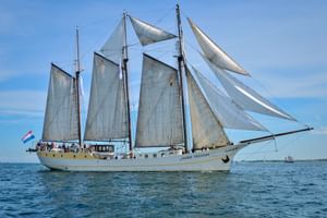 Three-masted sailing ship Mare Frisium with white sails unfurled on calm blue water under a clear sky. Dutch flag visible at stern.