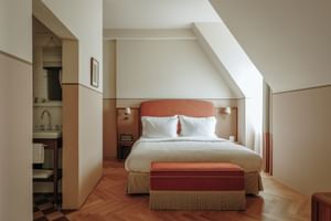 Modern double room with coral-colored headboard, white bedding, herringbone parquet flooring, and sloped ceiling. Bathroom visible through doorway.