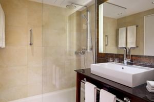 Modern hotel bathroom at Sheraton Essen with glass shower, white rectangular sink on dark countertop, large mirror, and beige tile walls.
