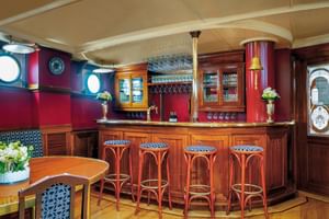 Wooden bar with four red bar stools on Mare Frisium ship. Red walls, portholes, hanging glasses, and decorative flowers create a cozy atmosphere.