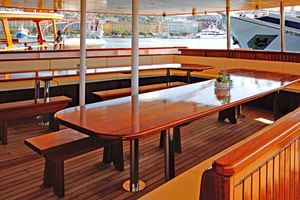 Wooden deck area on MS Aria with polished tables, benches, and curved seating. Large windows show harbor views with buildings and boats.