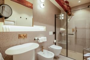 Modernes Bad mit Terrazzowänden, freistehender Badewanne, Glasdusche, Bidet und sichtbaren Holzbalken. Goldfarbene Armaturen und runde Wandleuchten.