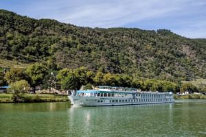 MS Switzerland river cruise ship sailing on calm green water with forested hills in the background under a partly cloudy sky.