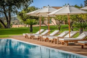Swimmingpool auf Finca Son Roig auf Mallorca mit weißen Sonnenliegen unter Sonnenschirmen auf Holzdeck, umgeben von Rasen und Palmen.