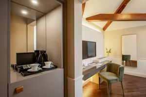 Hotelzimmer in der Villa Soligo mit Holzbalken, Schreibtisch mit grünem Stuhl, Wandfernseher, Spiegel und Kaffeestation mit Wasserkocher in Nische.