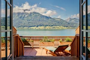 Wooden balcony with sun lounger overlooking a lake and forested mountains. Open balcony doors frame the scenic Austrian alpine landscape.