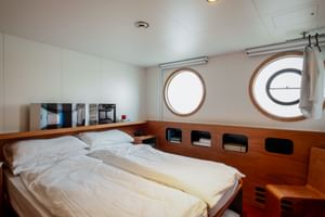 Schiffskabine auf der MS Ave Maria mit Doppelbett, weißer Bettwäsche, Holzverkleidung, zwei runden Bullaugen und eingebauten Staufächern.