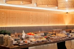 Breakfast buffet with fresh fruit, bread, cereals and pastries on wooden table. Modern dining room with wicker lamps and wood paneling.