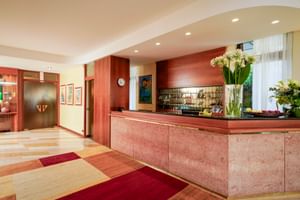 Modern hotel reception with pink marble counter, wooden paneling, and checkered floor in warm tones. White orchids decorate the desk.
