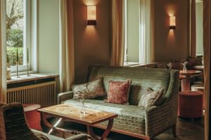 Cozy lounge at Parkhotel Mondschein Bozen with green sofa, wooden coffee table, wall sconces, and large windows overlooking garden.