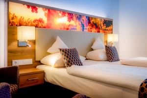 Modernes Doppelzimmer mit zwei Einzelbetten, farbig beleuchtetem Kopfteil, weißen Kissen, gemusterten Zierkissen und Holznachttischen mit Lampen.
